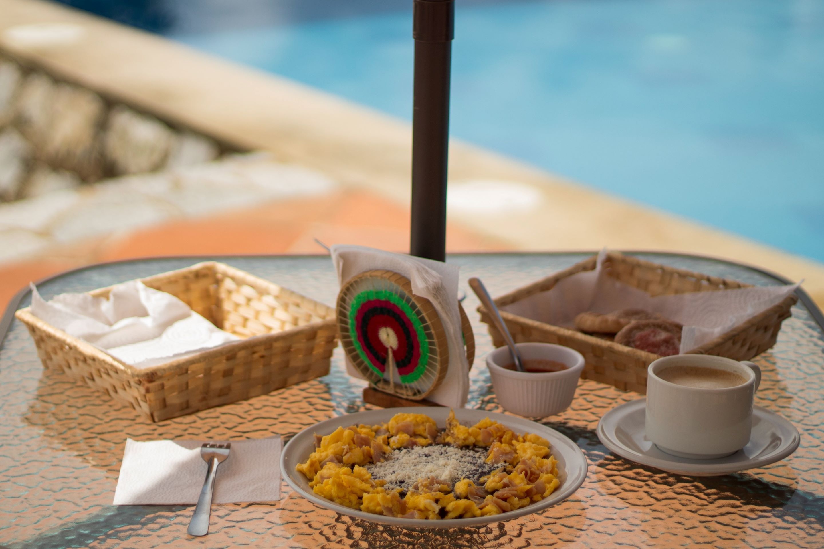 A table with a plate of scrambled eggs, a cup of coffee, and a basket of pastries by a poolside.