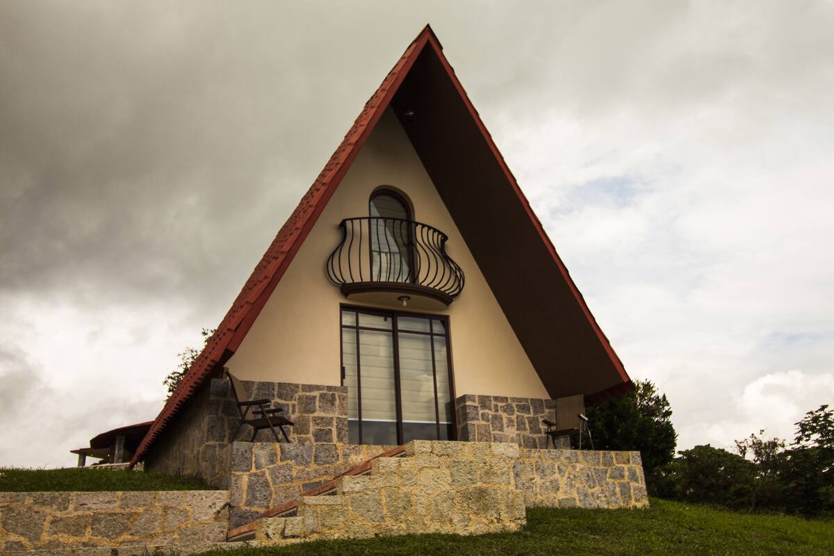 Un edificio triangular con base de piedra y techo rojo. Tiene un balcón con barandilla de metal y un ventanal grande.
