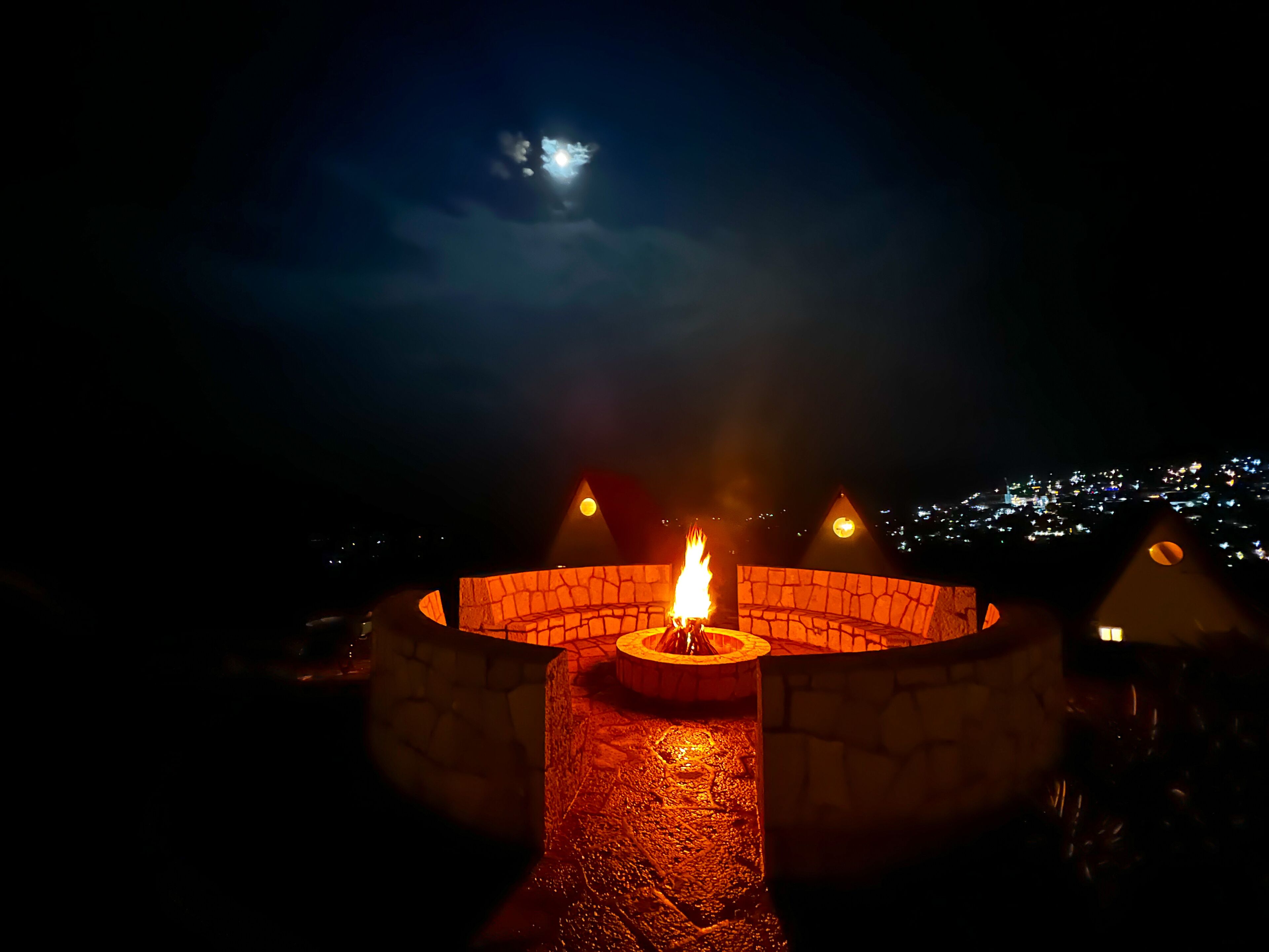 A circular stone fire pit with a central flame, surrounded by a stone wall, set against a night sky with a visible moon.