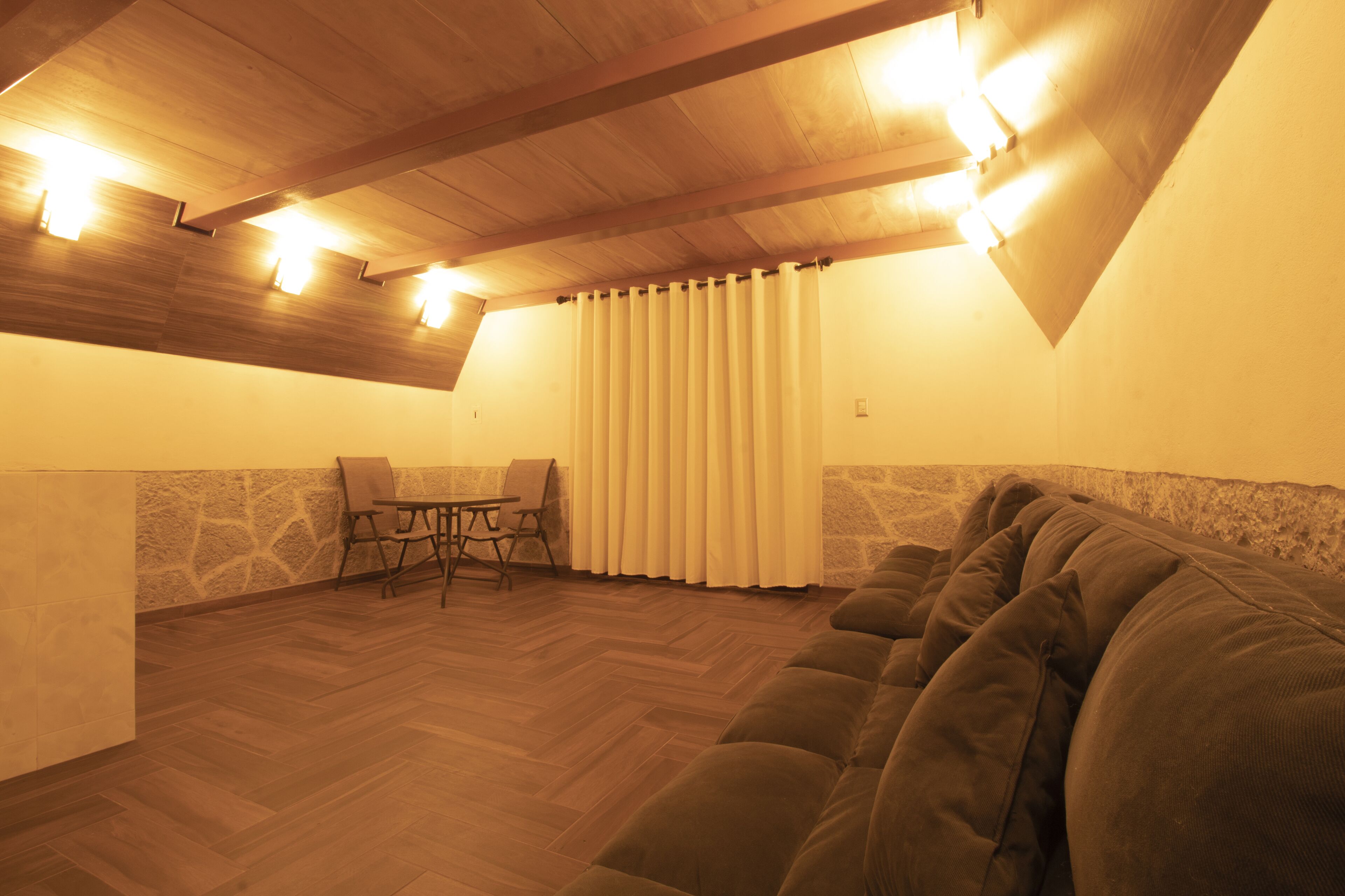 Un dormitorio acogedor en el desván, con techo de madera, un ventanal con cortinas, un sofá y una pequeña mesa con sillas.