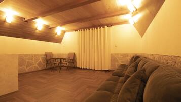 Een gezellig slaapkamer op zolder met houten plafond, een groot raam met gordijnen, een bank en een kleine tafel met stoelen.