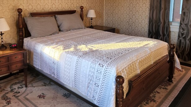 Un dormitorio con una cama grande, cabecera de madera, dos mesitas de noche con lámparas, una alfombra estampada con motivos florales y una ventana con cortinas.