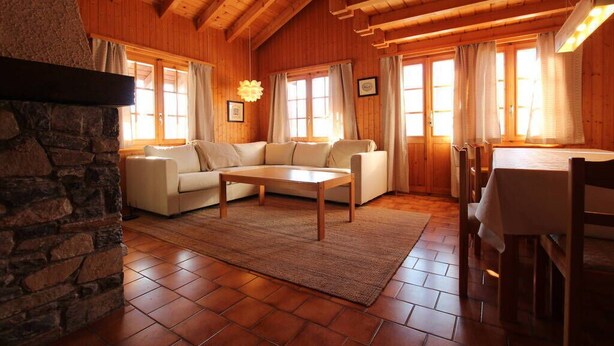 Una sala de estar acogedora con chimenea de piedra, vigas de madera en el techo, un conjunto de sofás y una mesa de comedor con sillas.