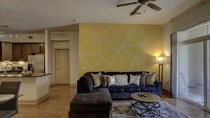 A modern living room with a sofa, ottoman, and coffee table, a kitchen in the background, and a large window with a view. - Luxury 2BD w/ pool. 5 min walk to NRG Stadium. 10 min Dr to Downtown Houston!! (Houston)