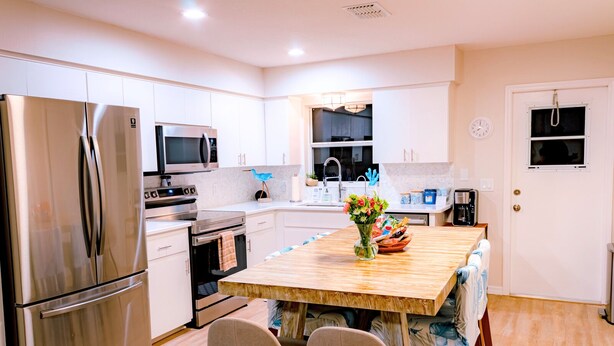 A modern kitchen with white cabinetry, stainless steel appliances, a wooden island with chairs, and a vase of flowers.