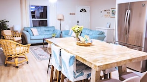 A modern living room with a blue sofa, a dining table with chairs, a ceiling fan, and a refrigerator.