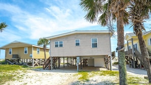 A beachfront house with a sandy beach in front, wooden deck, and a palm tree.