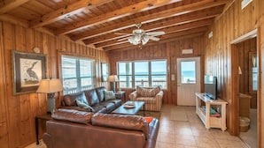 A wooden cabin interior with a leather sofa, a striped armchair, a ceiling fan, and a television on a small table.