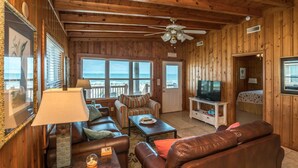 A cozy cabin interior with a wooden interior, a ceiling fan, a television, and a view of the ocean through large windows.
