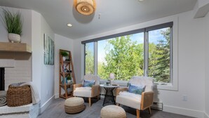 A modern living room with two white armchairs, a round ottoman, a fireplace, a large window with a view of trees, and a small table with a lamp. - Stunning & Modern Family-Friendly Slopeside Retreat in the Heart of Snowmass! (Snowmass Village)