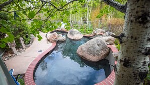 A circular hot tub surrounded by rocks and a brick border, with a pathway leading to a wooden deck. - Stunning & Modern Family-Friendly Slopeside Retreat in the Heart of Snowmass! (Snowmass Village)