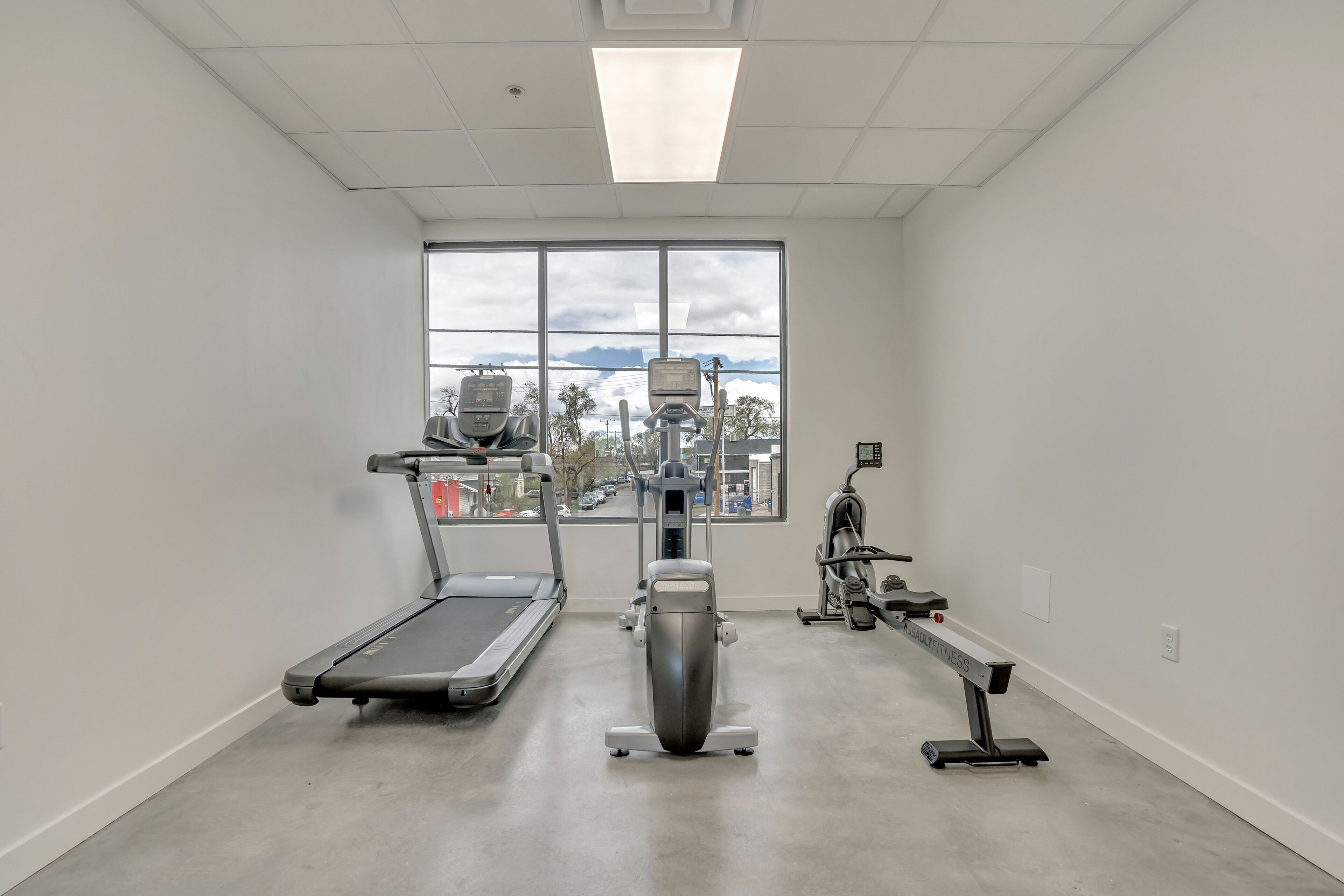 Un gimnasio con cinta de correr, elíptica y máquina de pesas, amplias ventanas con vista a la ciudad, y un diseño moderno y limpio.