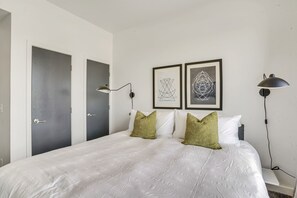 Cama bem arrumada com roupa de cama branca e dois travesseiros verdes, uma luminária de cabeceira e duas obras de arte abstratas emolduradas na parede.