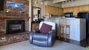 Un salon chaleureux avec une cheminée, un fauteuil en cuir et un coin cuisine doté de meubles en bois et d’un four à micro-ondes.