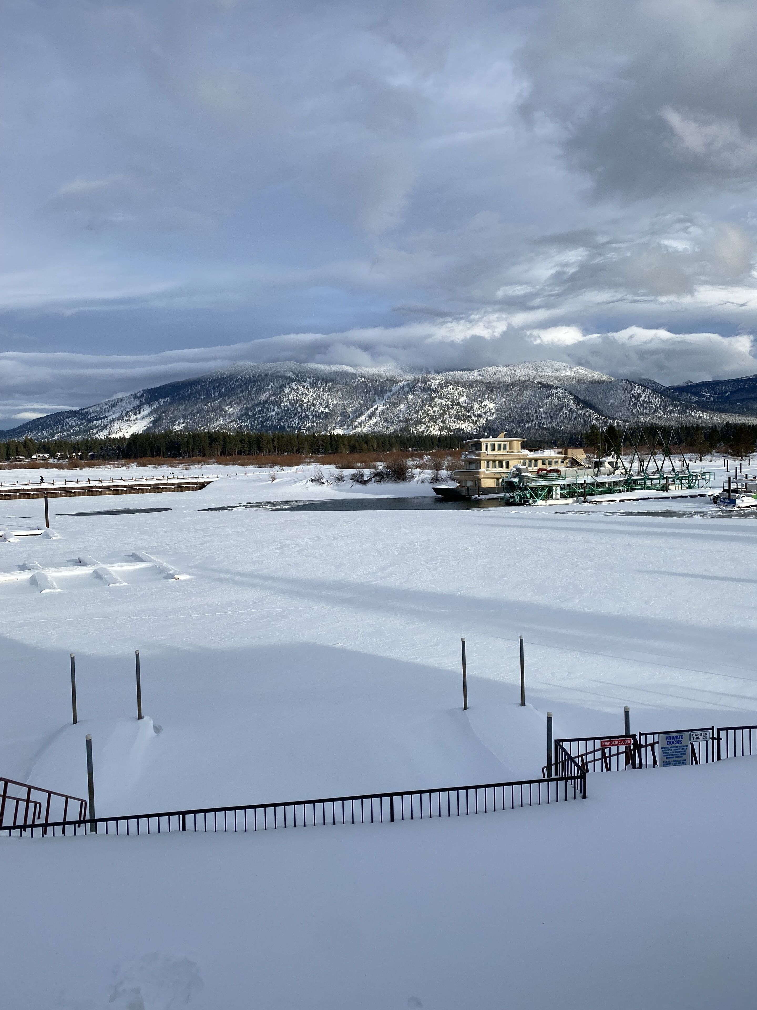 A snowy landscape with a mountain range in the background, a building with a balcony, and a fenced area in the foreground.