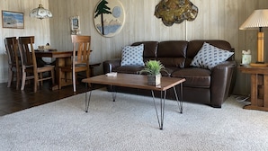 Un salon avec un canapé en cuir marron, une table basse en bois et un tapis blanc. On y trouve également un coin repas avec des chaises et une table en bois, ainsi qu’un tableau accroché au mur.