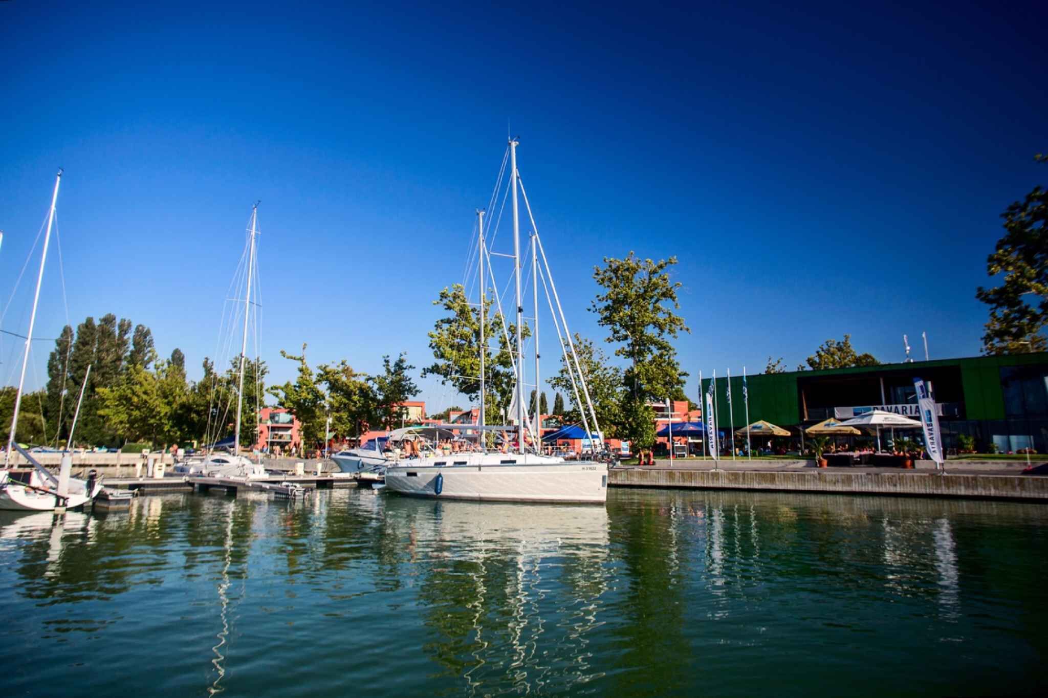 A marina with several sailboats docked, surrounded by trees and a building with outdoor seating.