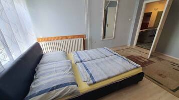Un dormitorio con cama, ventana con cortinas translúcidas, radiador y espejo en la pared.