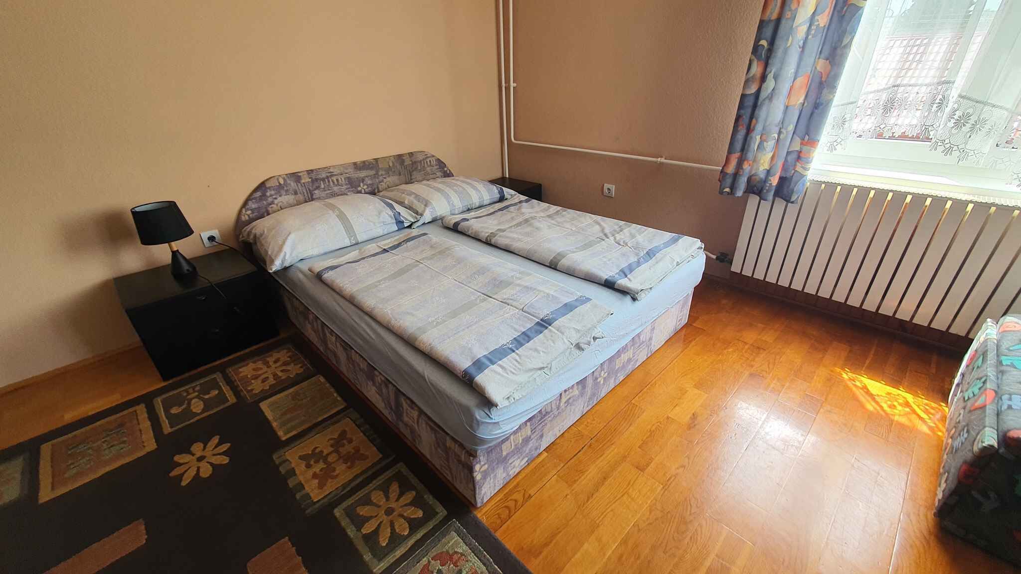 A bedroom with a bed, a nightstand, a lamp, a window with curtains, and a radiator.