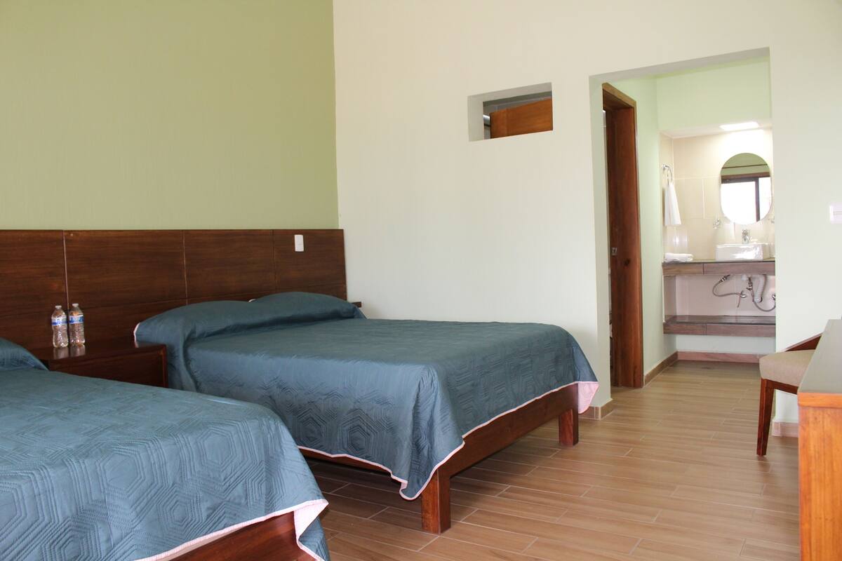 Habitación de hotel con dos camas, cabecero de madera, baño pequeño con espejo y una silla de madera.