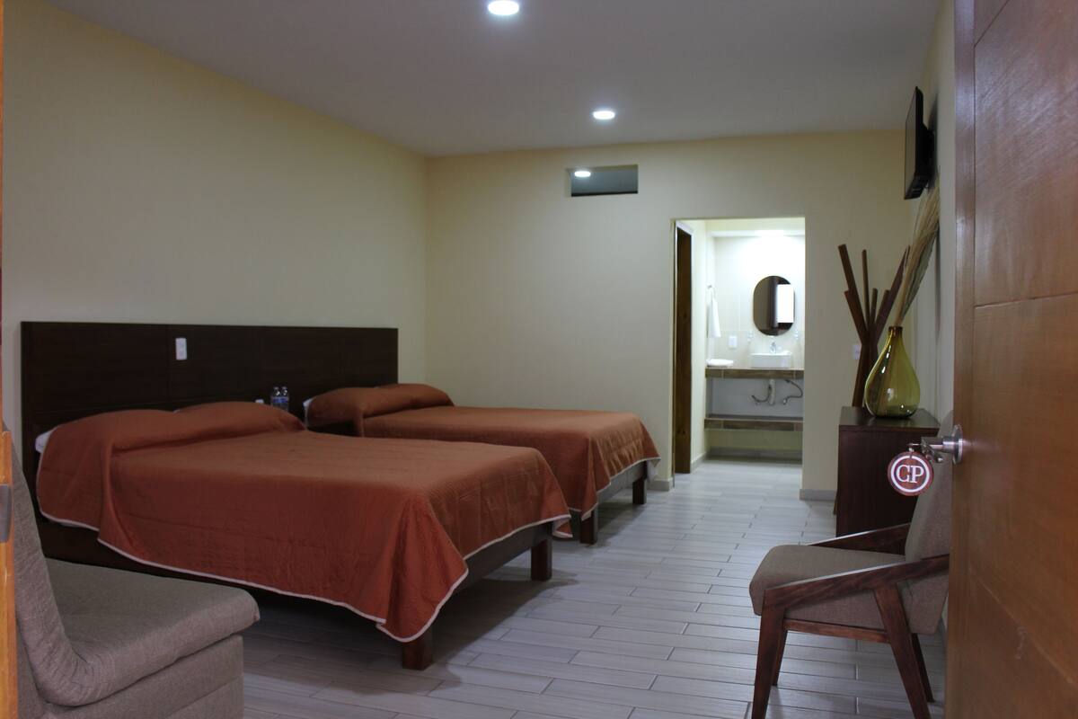 Habitación de hotel con dos camas, una silla, una mesita y un baño visible a través de una puerta abierta.