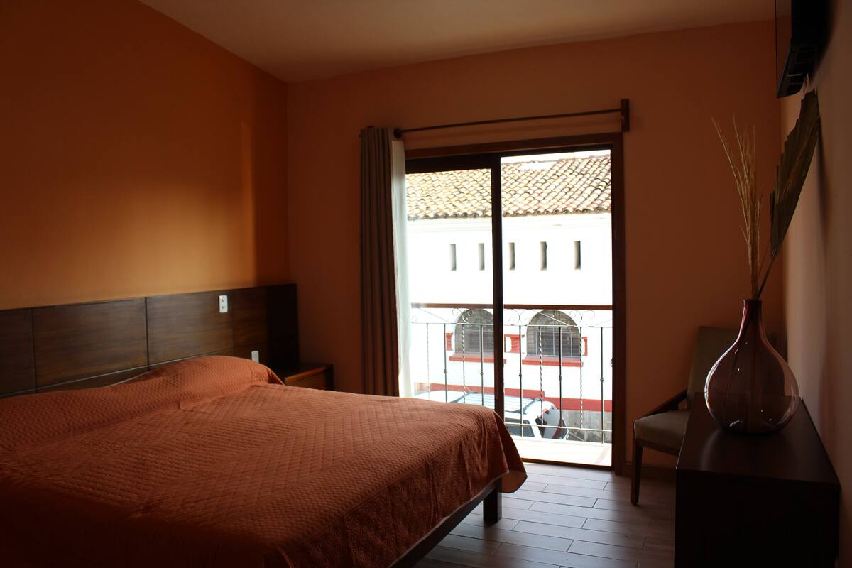 Habitación de hotel con cama, una silla, mesita de noche, un jarrón y vista al balcón exterior.