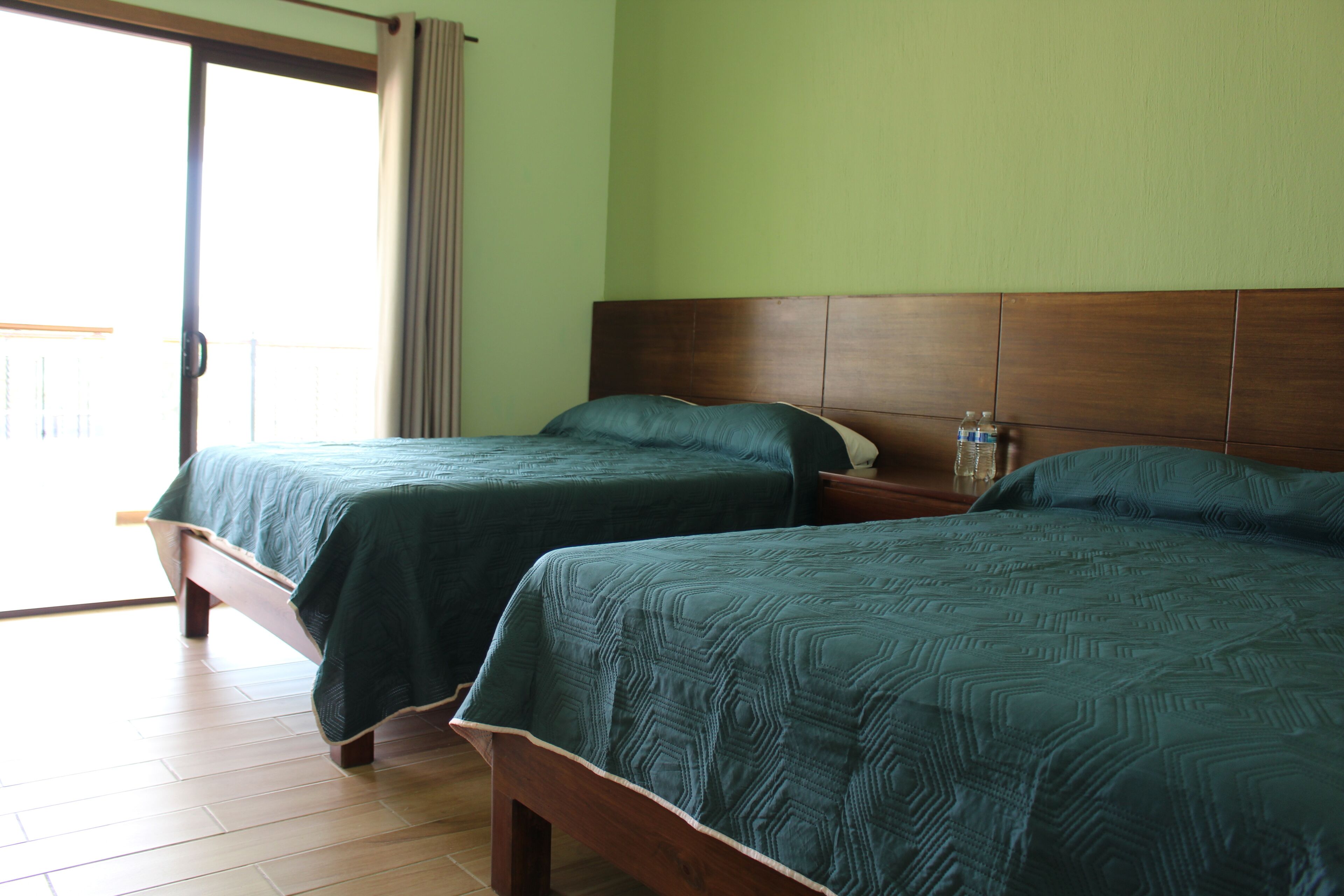 Dos camas individuales con cabeceros de madera en una habitación con pared verde y puerta corrediza de vidrio que da a un balcón.
