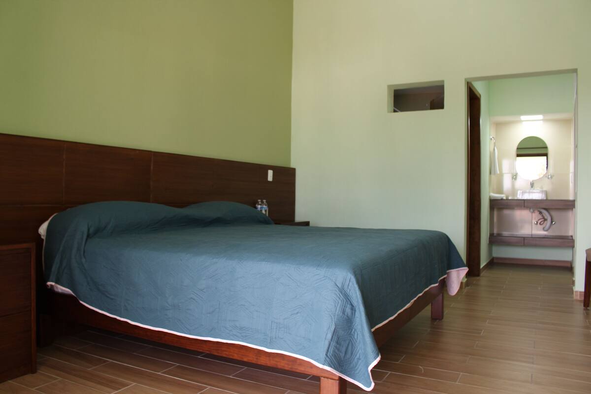 Un dormitorio con cama de madera, una mesita de noche, una pequeña tocador y un baño visible a través de una puerta abierta.