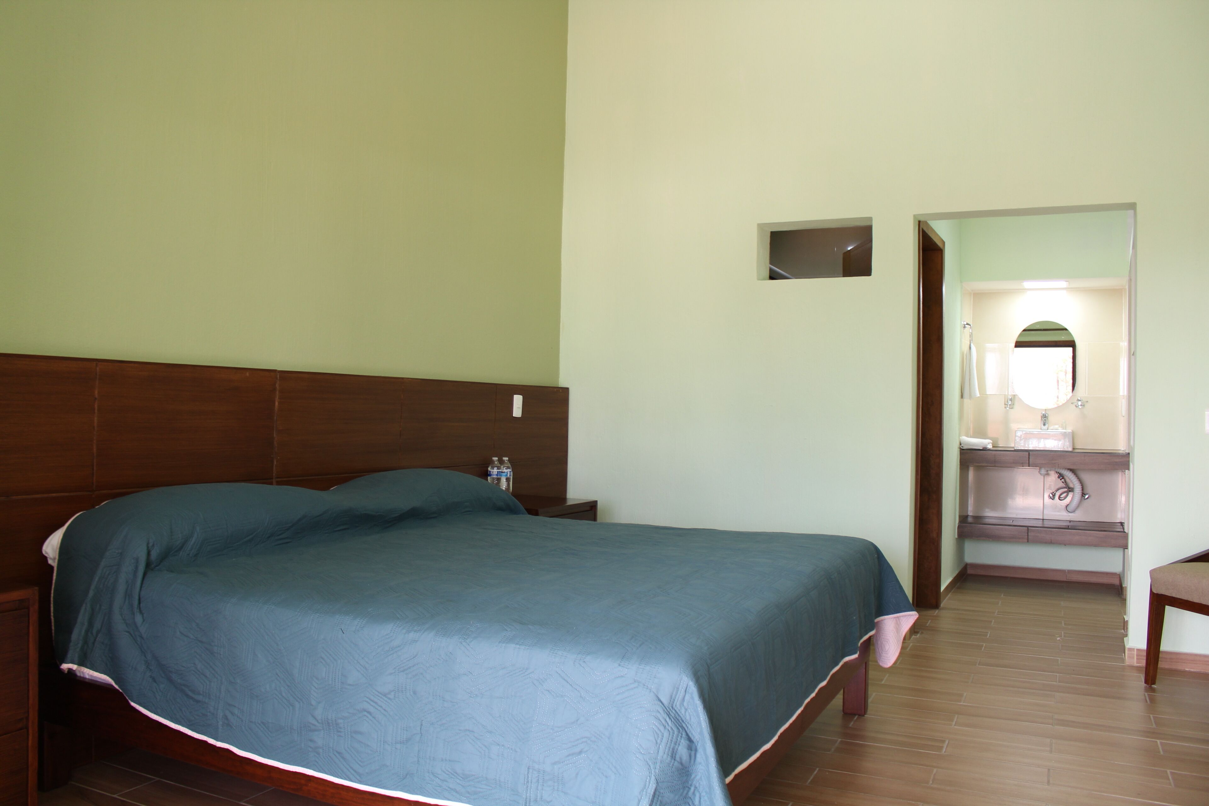 A bedroom with a large bed, a nightstand, a wooden headboard, a small mirror, and a bathroom visible through an open door.
