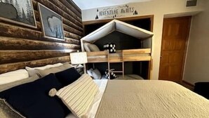 A bedroom with a bunk bed, a bed with a striped blanket, a wall-mounted shelf with shoes, and a 'Adventure Awaits' sign. - Heart of the mountain/5 Beds/Peaceful location with large deck (Gatlinburg)