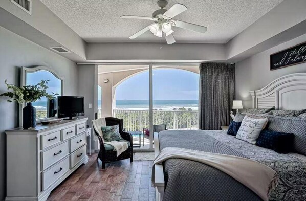 A bedroom with a balcony, a ceiling fan, a TV, a dresser, and a bed with patterned bedding.