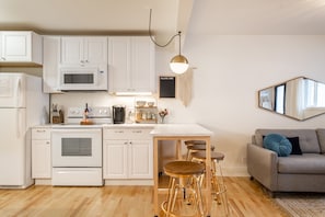 Una cocina moderna con armarios blancos, microondas integrado y cafetera. Hay una mesa de comedor de madera con taburetes y un sofá gris con un cojín azul.