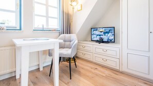 Ein Zimmer mit weißem Schreibtisch, einem Sessel, einem Fernseher auf einer Kommode und einem Fenster mit Vorhängen.