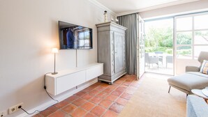 Ein Hotelzimmer mit einem an der Wand montierten Flachbildfernseher, einem weißen Schminktisch, einem grauen Schrank, einem grauen Sofa und einer großen Glasschiebetür, die zu einem Außenbereich mit einer Terrasse führt.