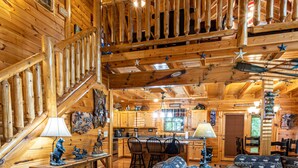 Interior de una cabaña de troncos con escalera de madera, sala de estar con sofá y cojines estampados, zona de cocina y mesa de comedor con sillas.