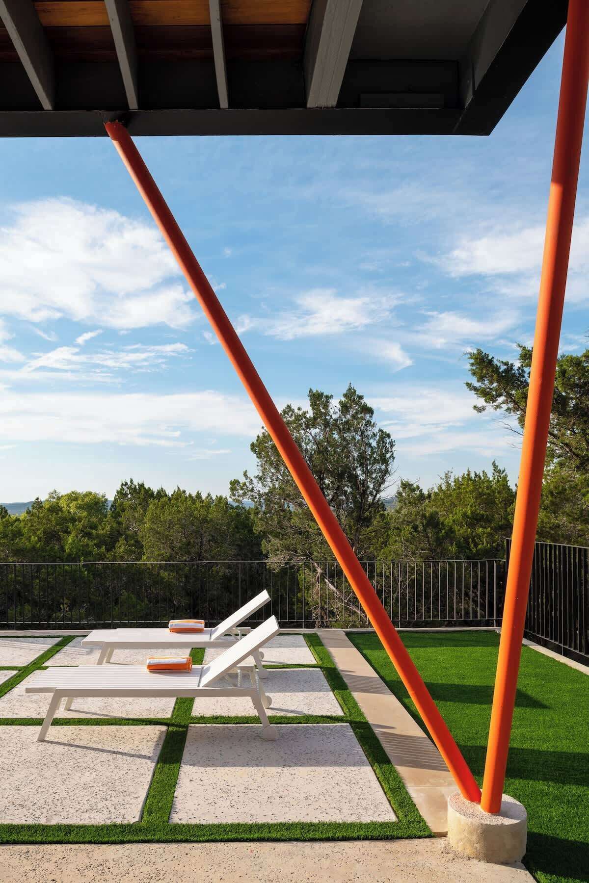 An outdoor seating area with white lounge chairs and orange cushions, surrounded by artificial grass and concrete flooring, under a wooden roof structure.