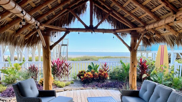 Un área de estar cubierta al aire libre con mobiliario de mimbre y una mesa, con vistas a una playa.