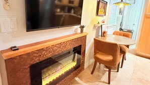 A modern living room with a flat-screen TV mounted on the wall, a built-in fireplace, a dining area with a table and chairs, and a patterned rug on the floor.