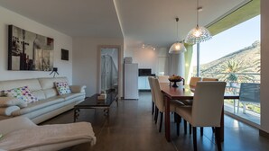 A modern living room with a sofa, dining table, and chairs, a kitchen in the background, and a large window with a view of a mountainous landscape. - VILLA OASIS - APART N - 5 (TELDE)
