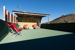 Un área de estar al aire libre con techado, sillas y mesas, un pequeño edificio con techo inclinado, y una colina al fondo.