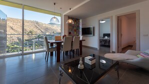 A modern living room with a dining area, large windows, and a view of a hilly landscape. - VILLA OASIS - APART N - 5 (TELDE)
