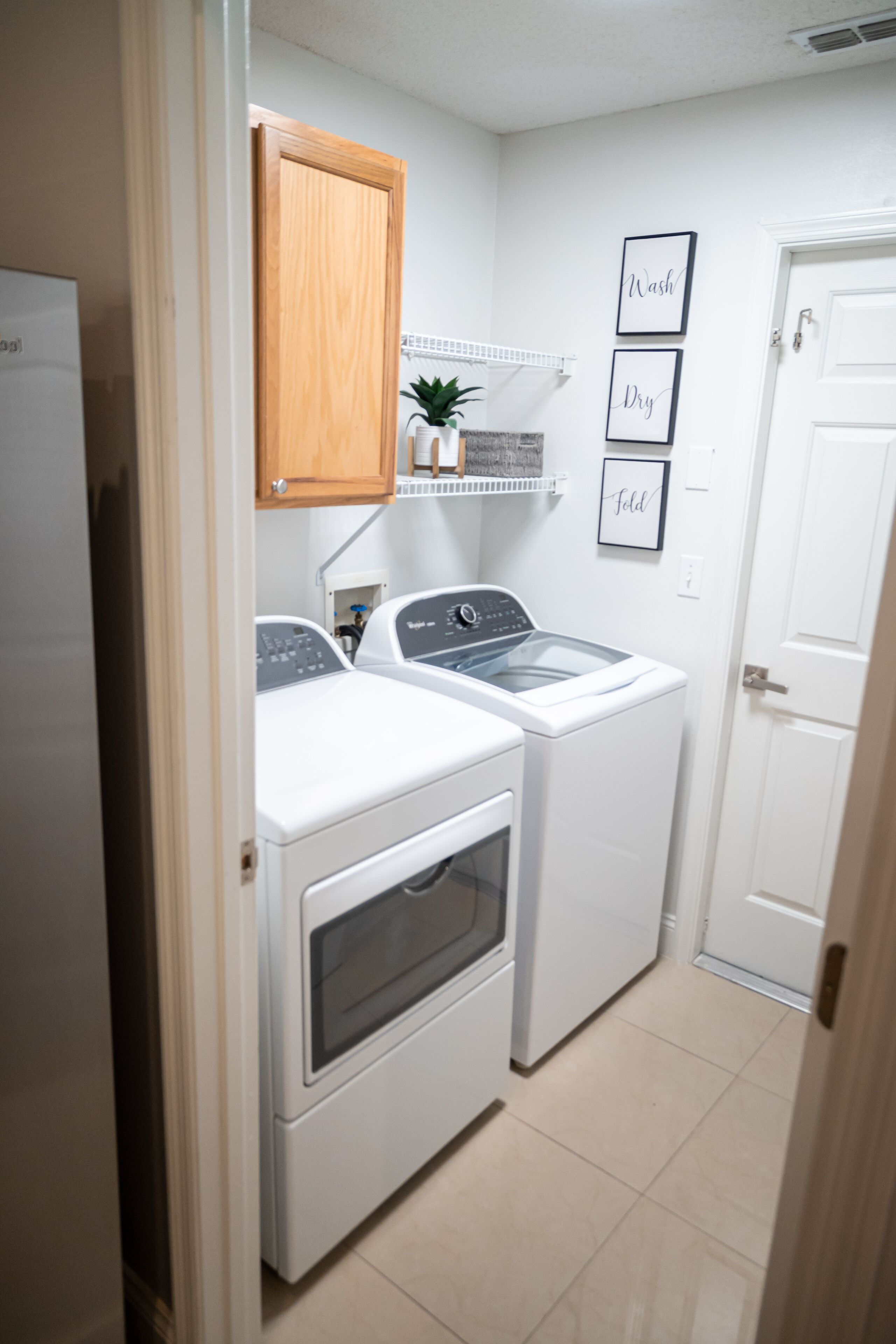 A laundry room with a washing machine, dryer, wooden cabinet, and a shelf with a plant.