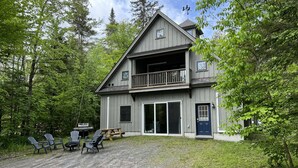A two-story cabin with a balcony, a covered patio with chairs, and a gravel driveway. - Sunapee Barn Retreat (Sunapee)