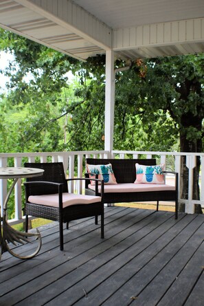 A covered porch with a wicker sofa and a small table, surrounded by white railings and lush greenery in the background. - New! Beautifully updated private home near White River and Bull Shoals (Flippin)