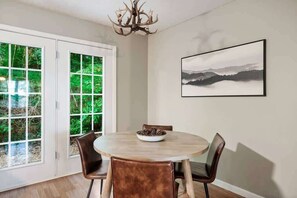 A dining area with a round table, four chairs, a chandelier, a framed landscape painting, and a view through large glass doors.