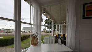 Una habitación con una mesa y silla blancas, una silla de mimbre, una cesta de mimbre con flores, y vistas a una calle y edificios.