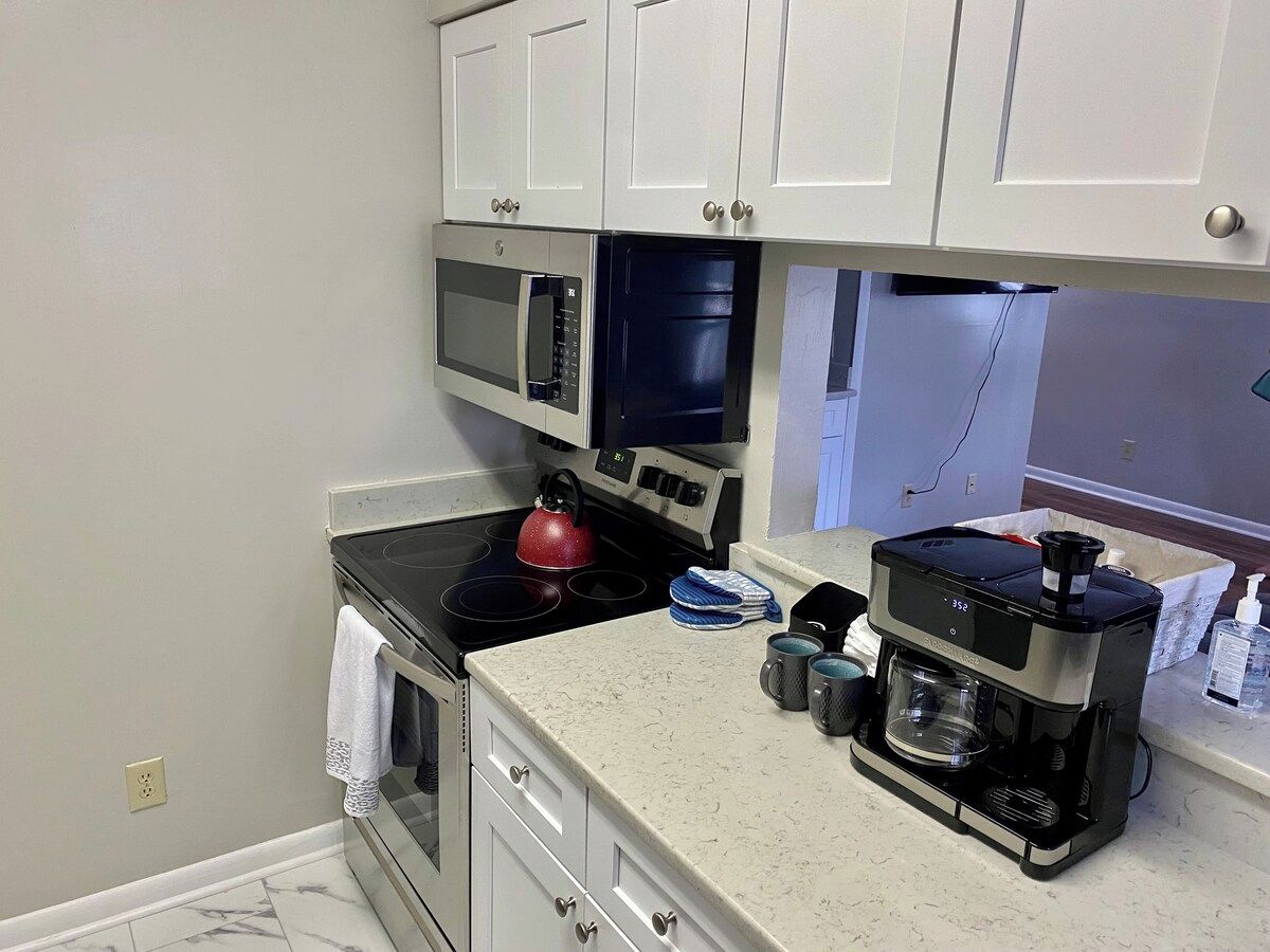 A kitchen with a microwave, oven, kettle, coffee maker, and a countertop with a kettle and mugs.