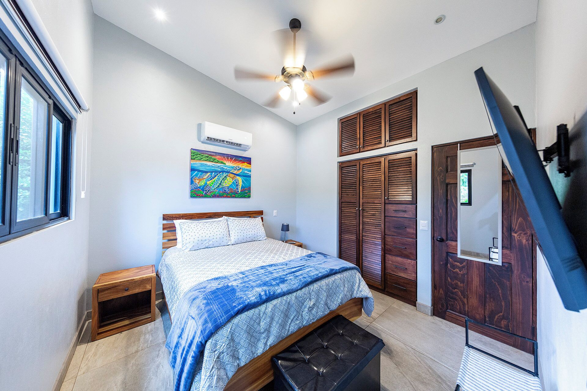 A bedroom with a bed, a ceiling fan, a window, a painting, and a wardrobe.