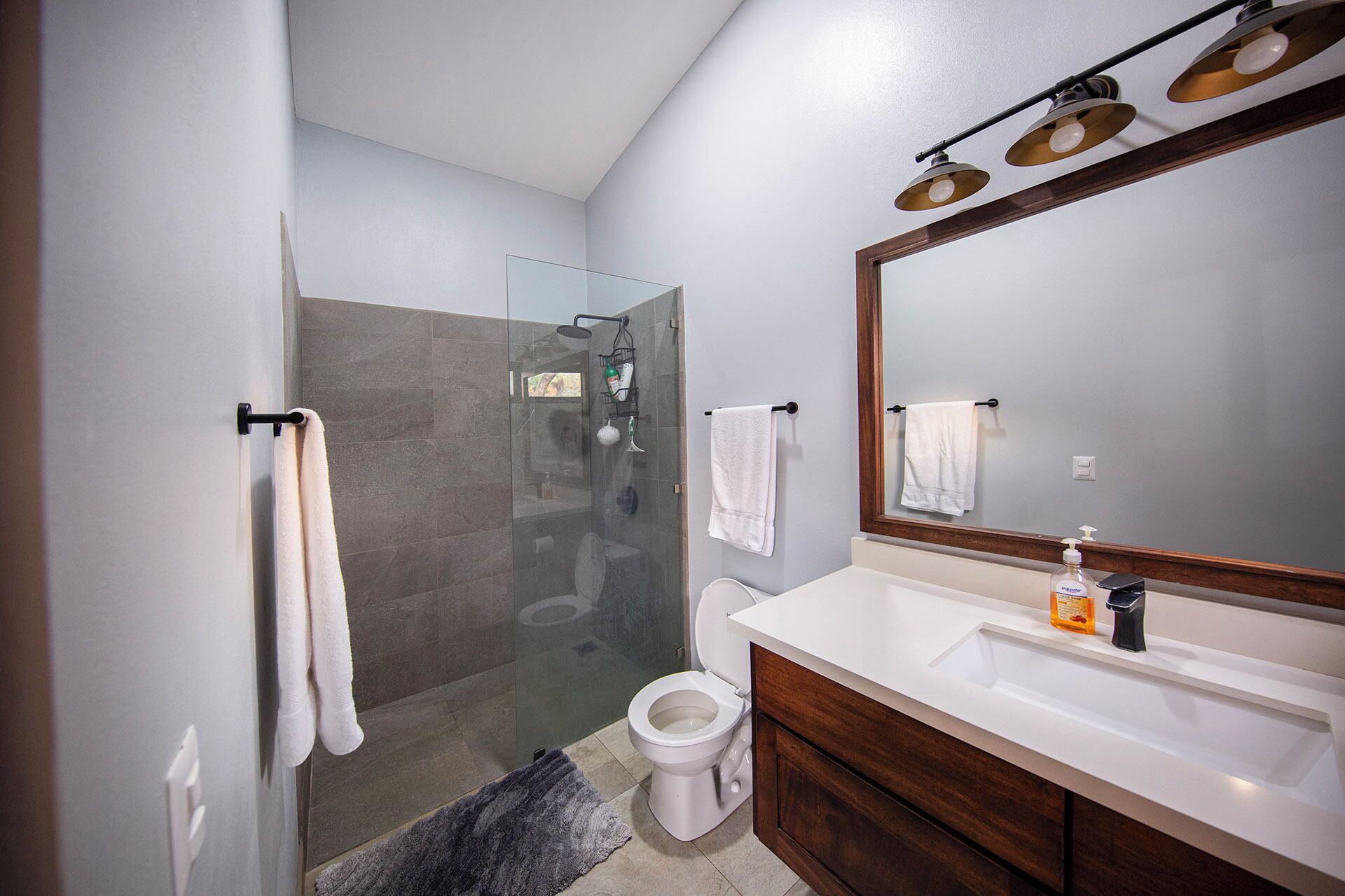 Un baño moderno con cabina de ducha de vidrio, inodoro, lavabo con espejo y dos toallas colgadas en ganchos.