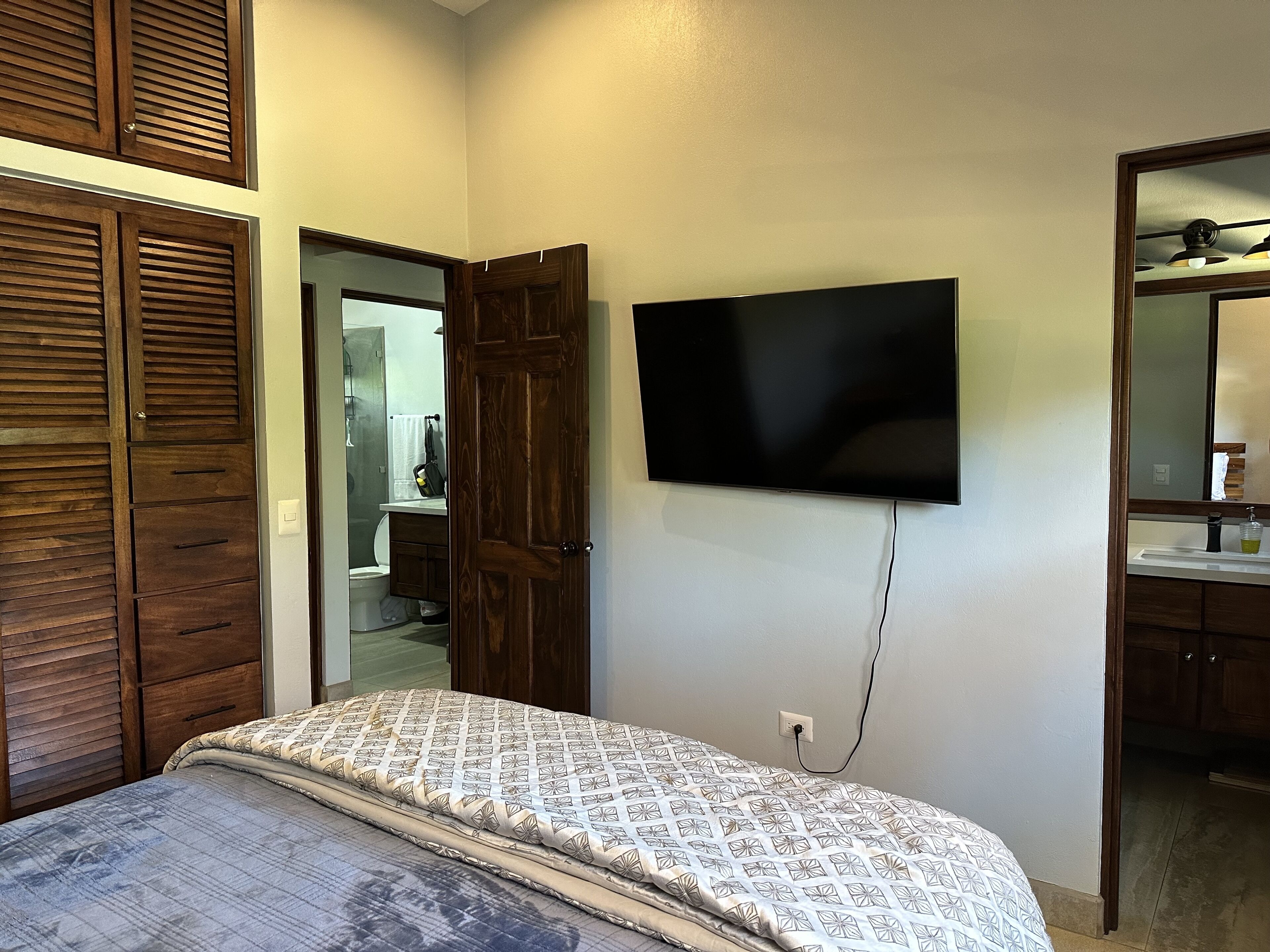 Een slaapkamer met een bed, een aan de muur bevestigde televisie, houten kasten en een badkamer zichtbaar door een geopende deur.
